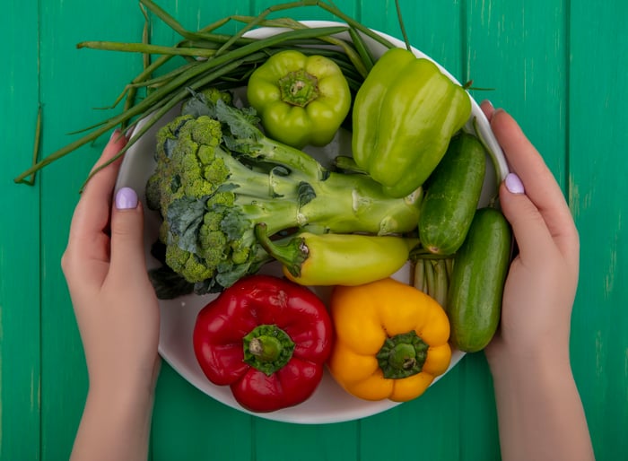 top-view-woman-holding-broccoli-with-cucumbers-colored-bell-peppers-green-onions-plate-green-background